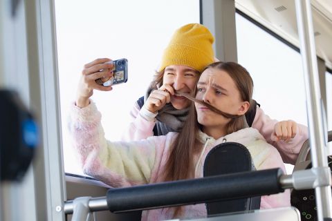 Kaksi nuorta istuvat bussissa, leikkivät toisen hiuksilla ja ottavat selfietä.