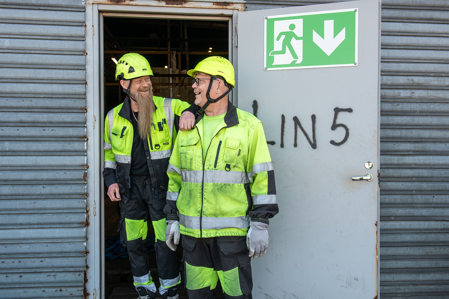 Kaksi henkilöä hymyilevät toisilleen suojavarusteissa.